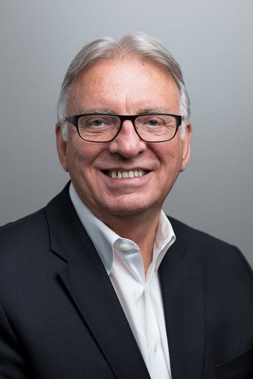 Man in suit smiling, wearing glasses; neutral grey background.