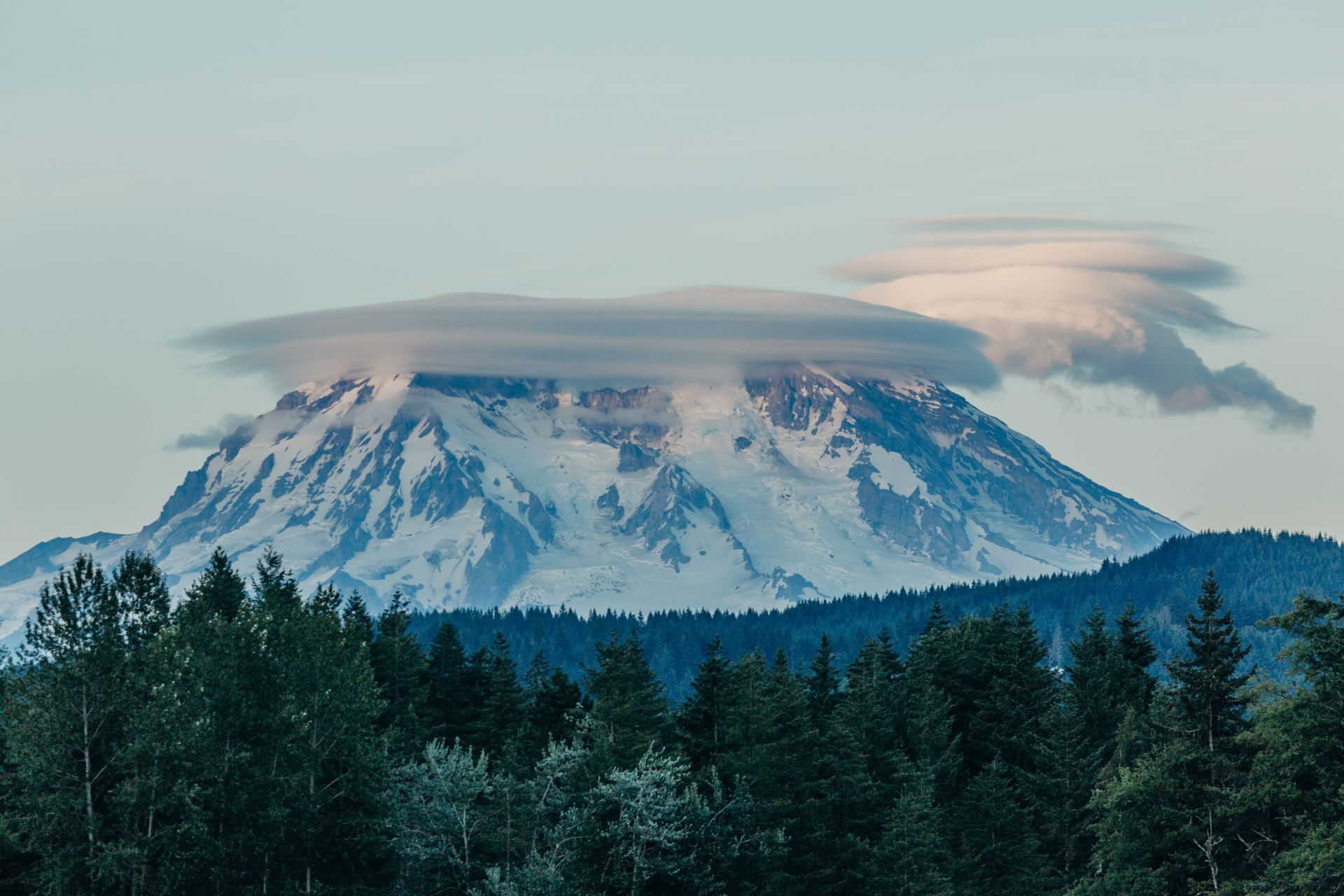 Snow-capped mountain obscured by a lenticular cloud, above a forest.