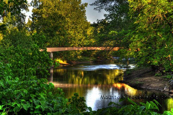 A calm river under a small bridge, surrounded by lush green trees and reflected sunlight.