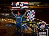 Driver in blue suit celebrates atop a dirt race car with trophy and checkered flag at night.