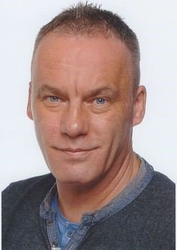 Ein Mann in einem grauen Pullover und einem blauen Hemd lächelt in die Kamera.