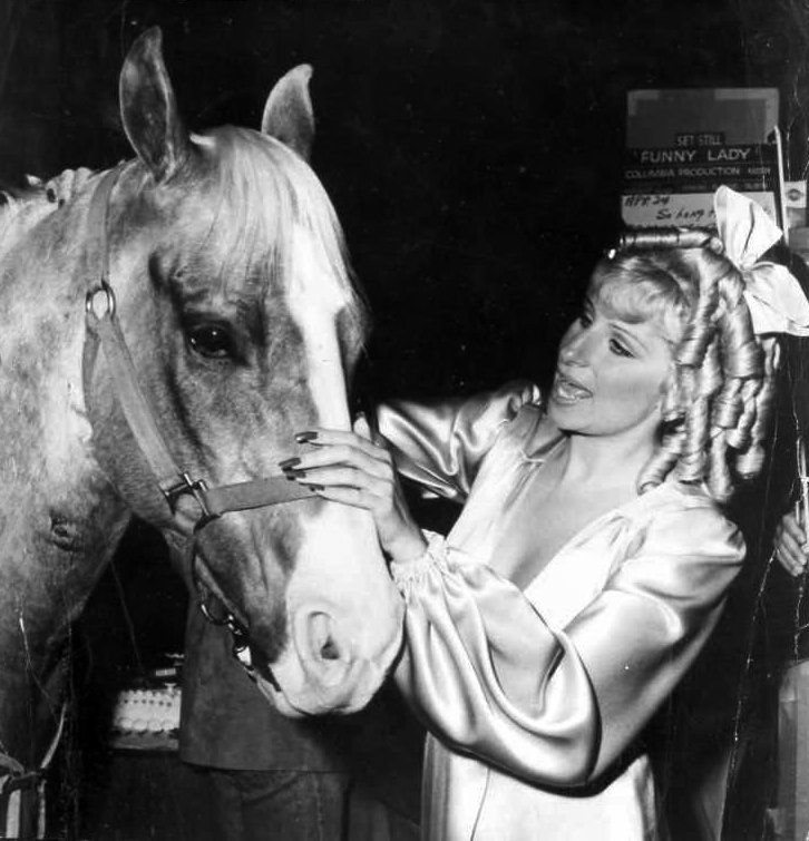 Barbra in satin dress and blond wig adjusting a horse’s bridle beside a poster in the background