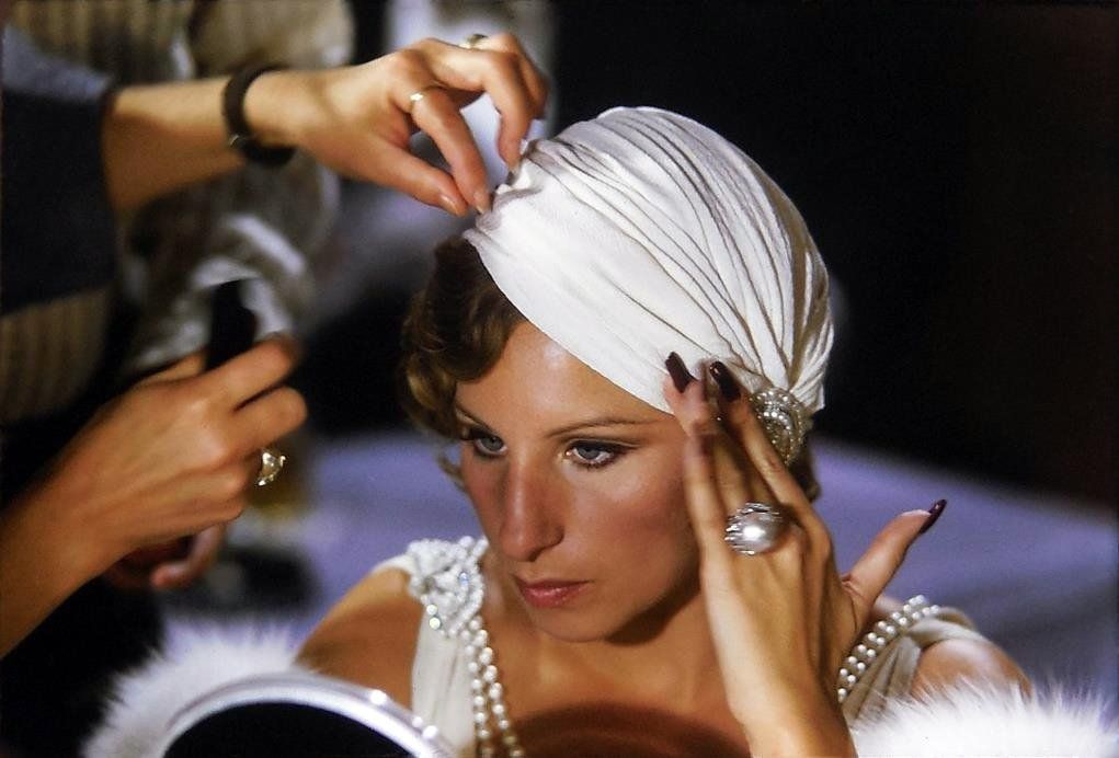 Barbra Streisand adjusting a white headwrap in front of a mirror as hands help style her hair