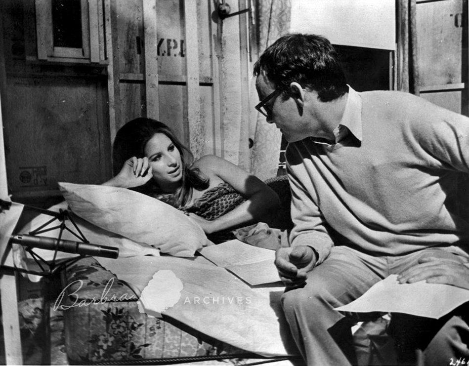 Barbra, reclined in bed on the set, speaks with the screenwriter, Buck Henry, who wears glasses.