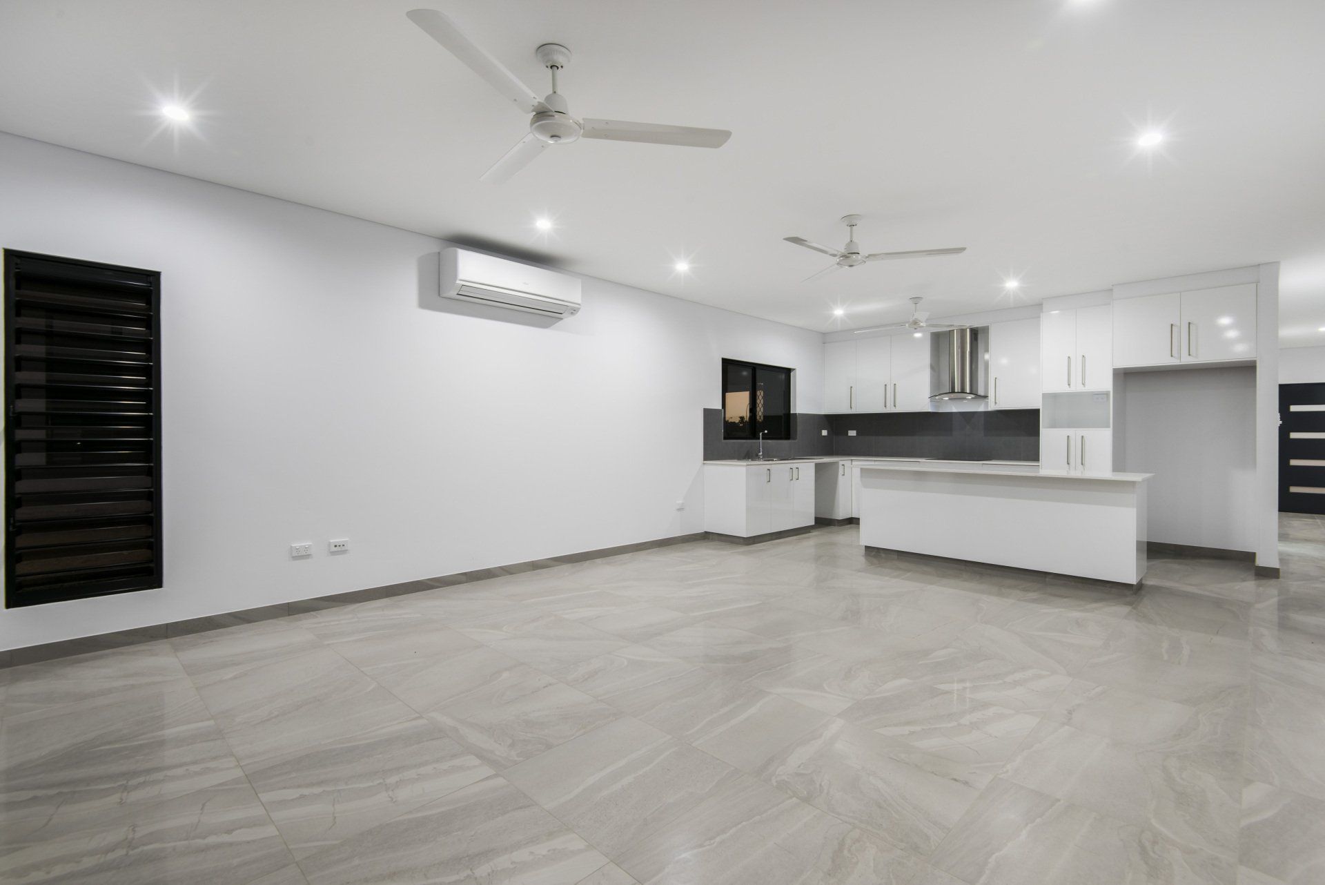 A large empty room with a ceiling fan and a kitchen in the background.