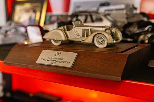 Silver car model on wooden base with plaque. Red and white background, other objects in the background.