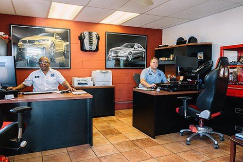 Two men in auto shop office, behind desks, looking at camera. Artwork of cars on wall.