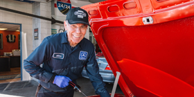 Mechanic smiling, working on a red car. He wears a blue uniform, cap, and gloves in a garage setting.