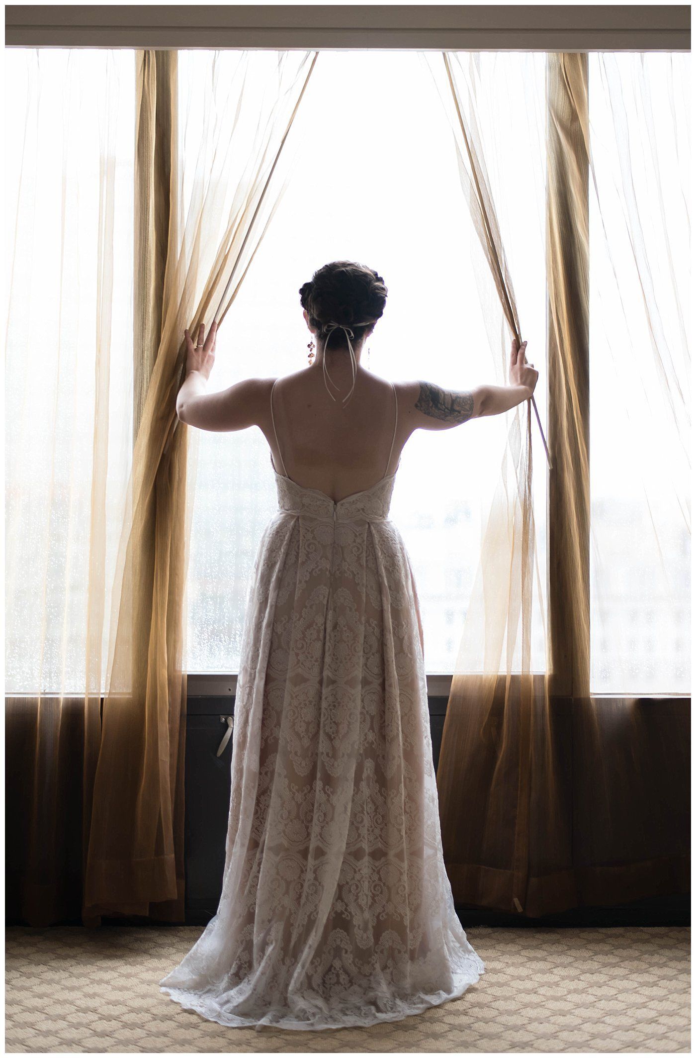 Woman in a long lace dress standing at a window, holding open sheer curtains with sunlight behind her