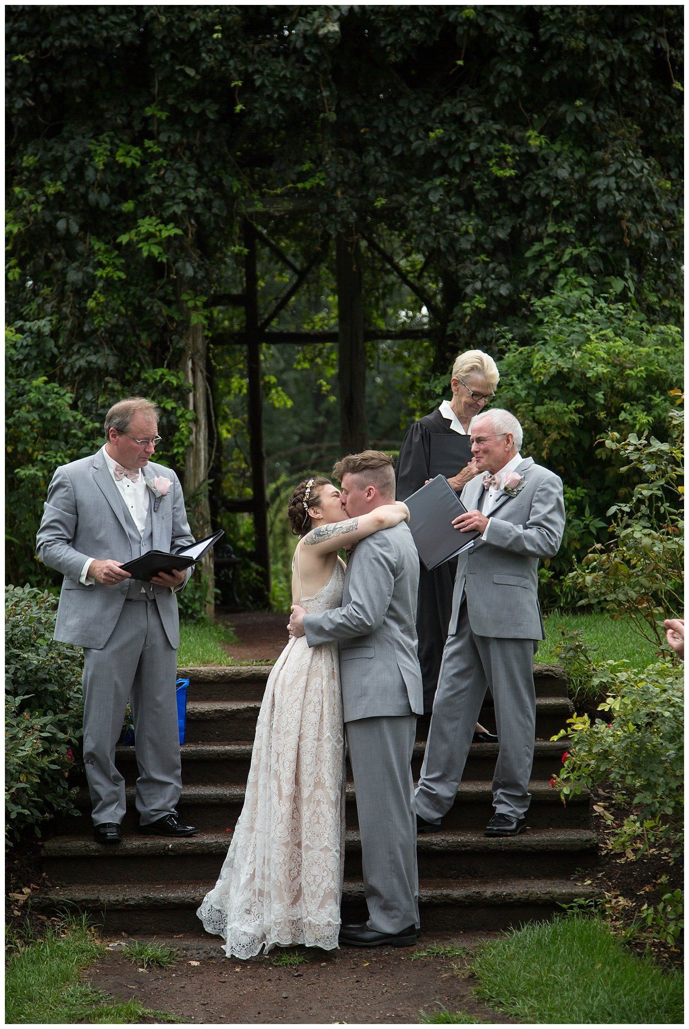 Outdoor wedding ceremony with couple embracing on steps, officiant reading nearby, and two guests behind them