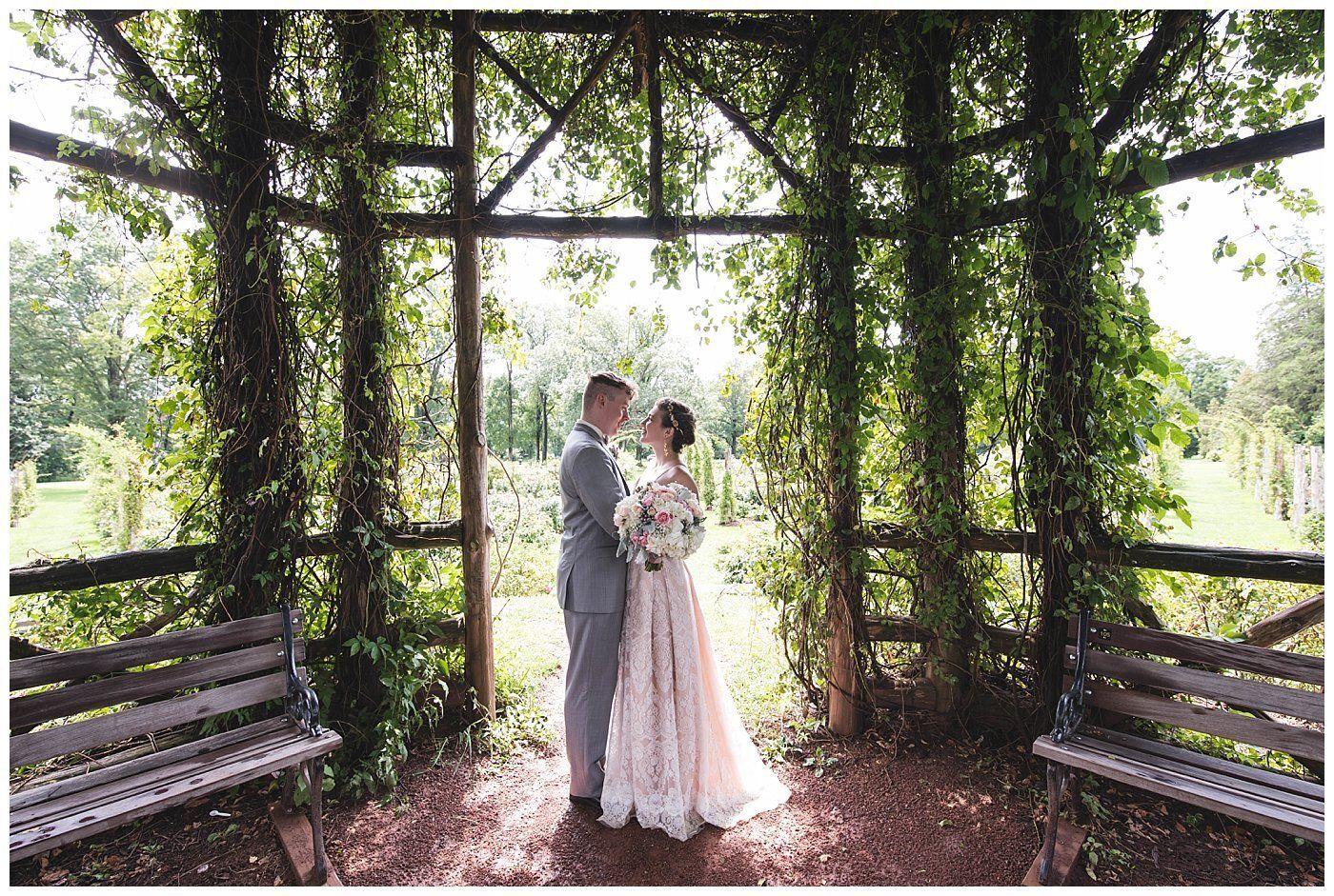 Couple in formal attire stands beneath a vine-covered gazebo, embracing in a sunlit garden.