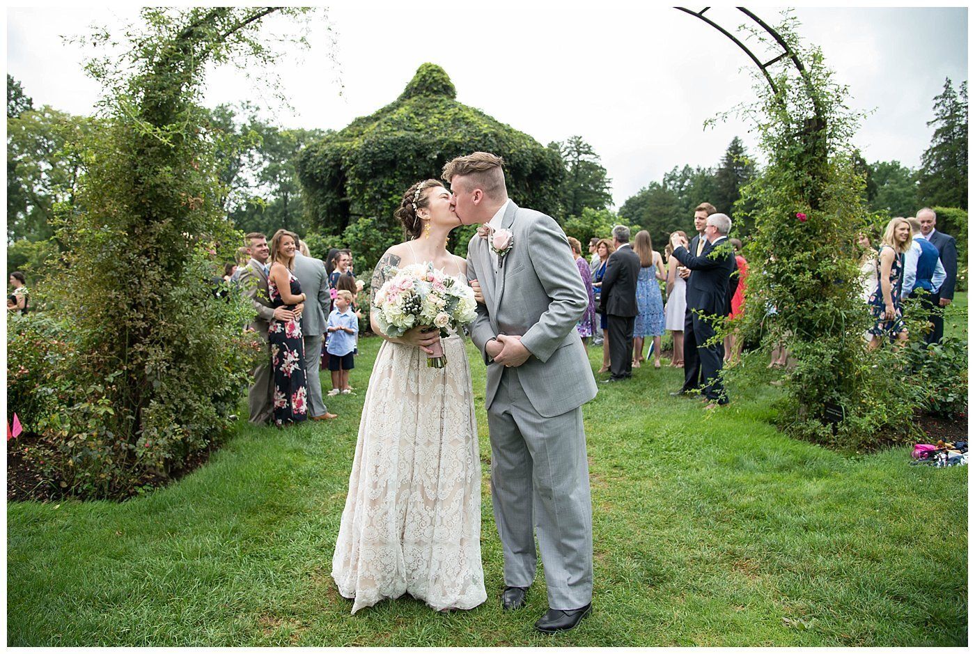 Newlyweds kiss in a garden wedding aisle surrounded by guests and greenery.