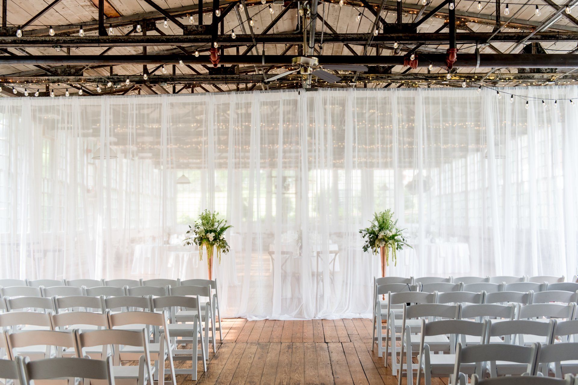 Rows of white chairs facing a sheer white curtain backdrop with two flower arrangements on a wooden floor.