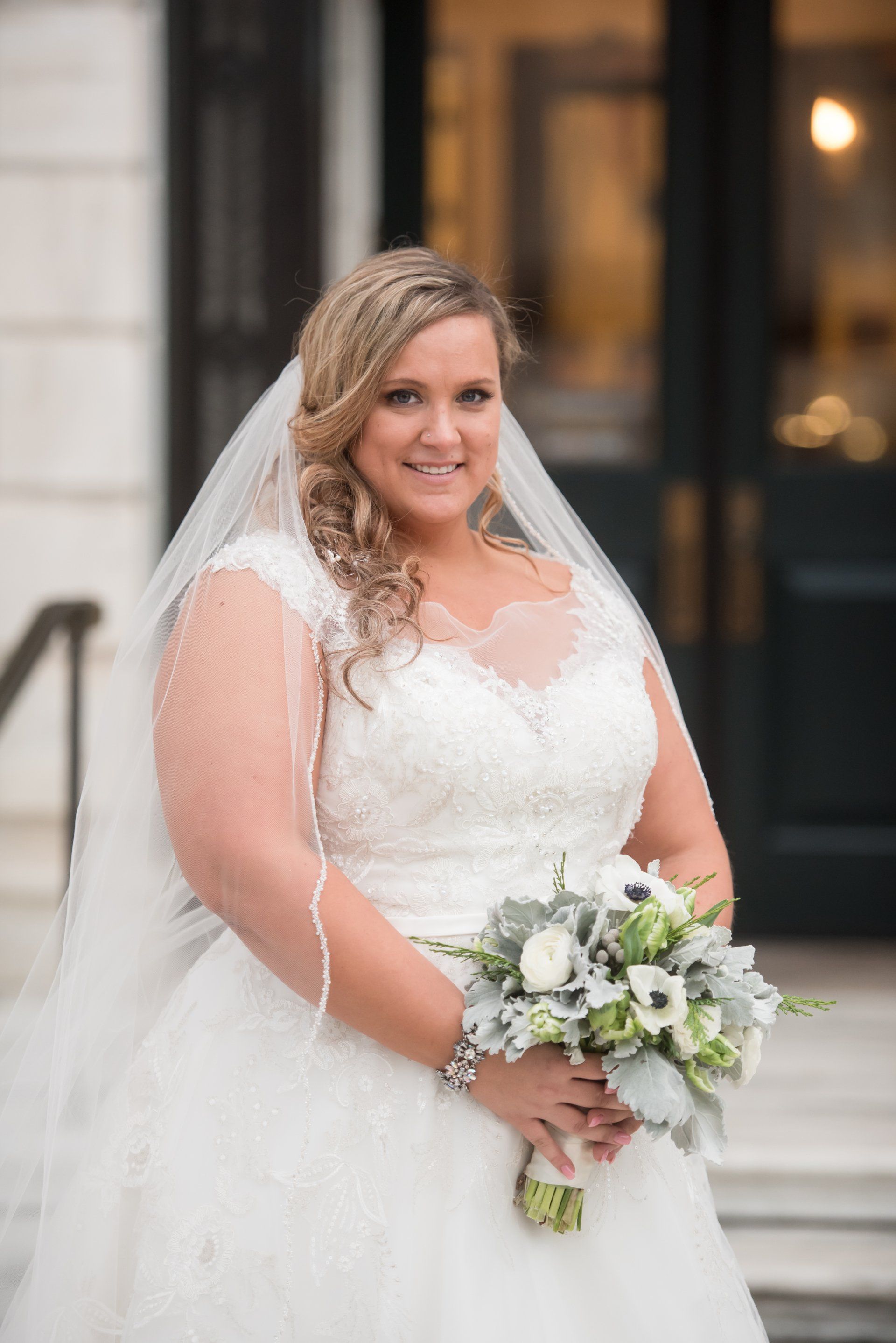 Bride in white lace gown and veil holding a bouquet, standing on steps outside a building.