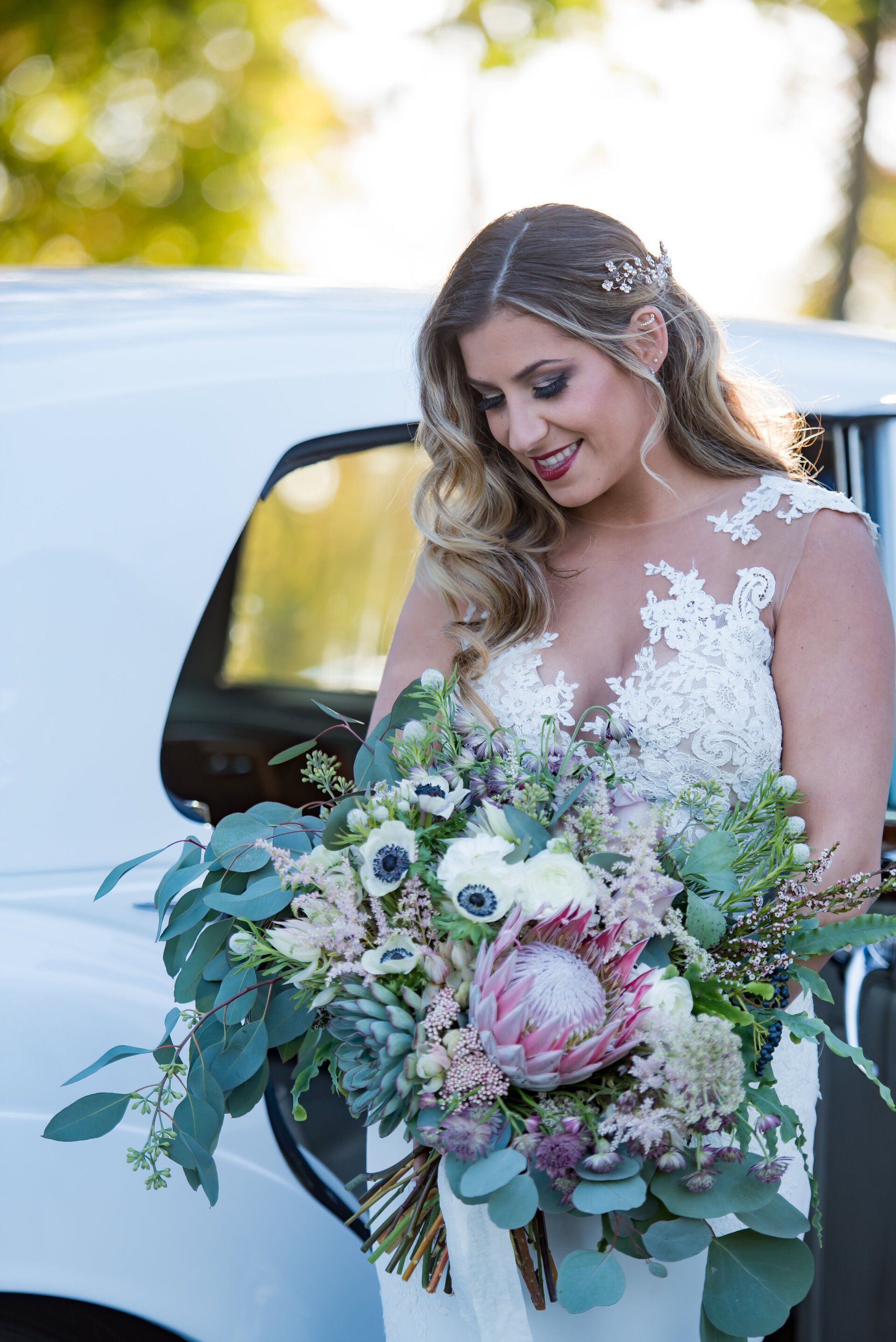Bride in a white lace gown holds a large pastel bouquet beside a white car outdoors.