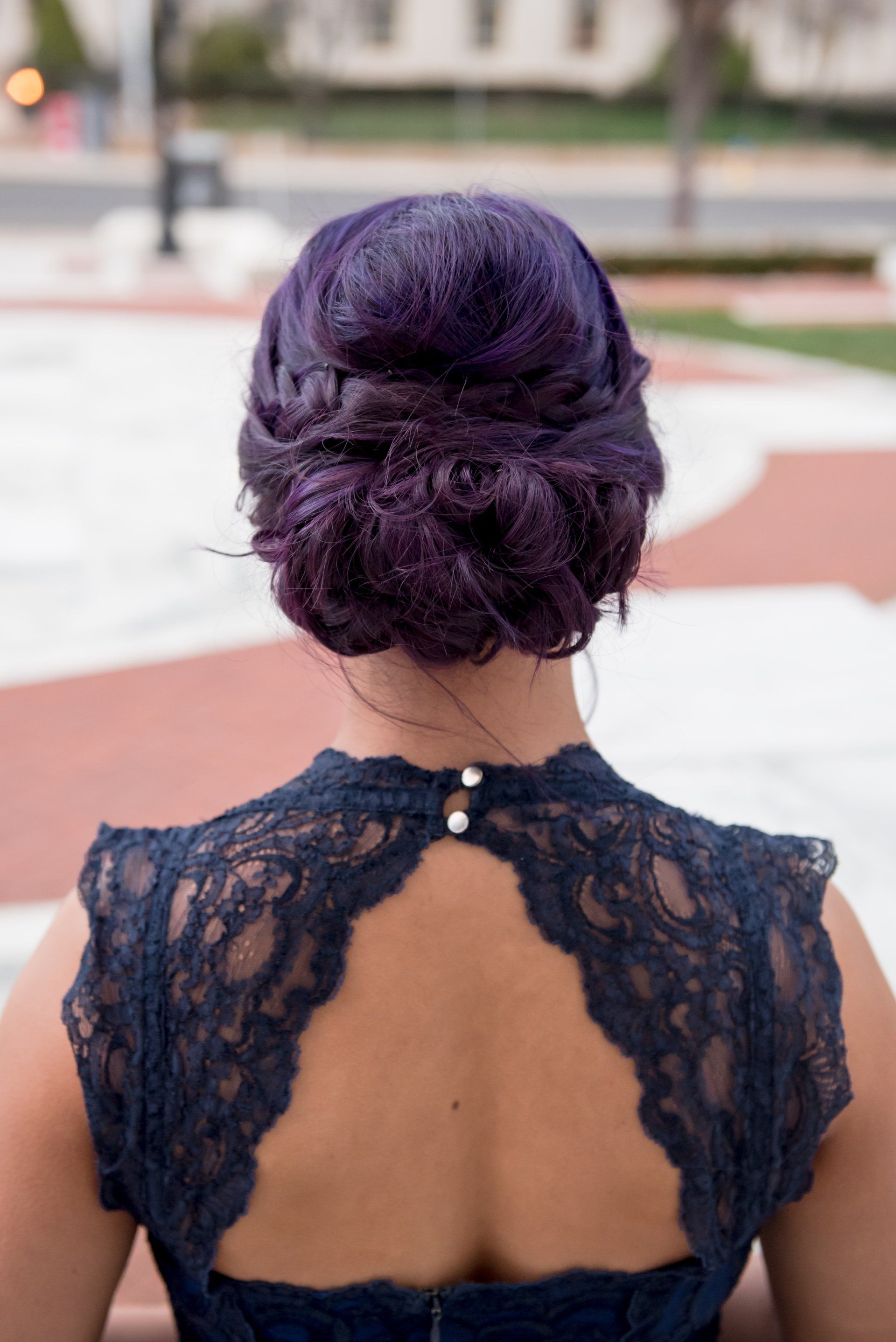 Woman with purple updo in a black lace dress, seen from behind outdoors