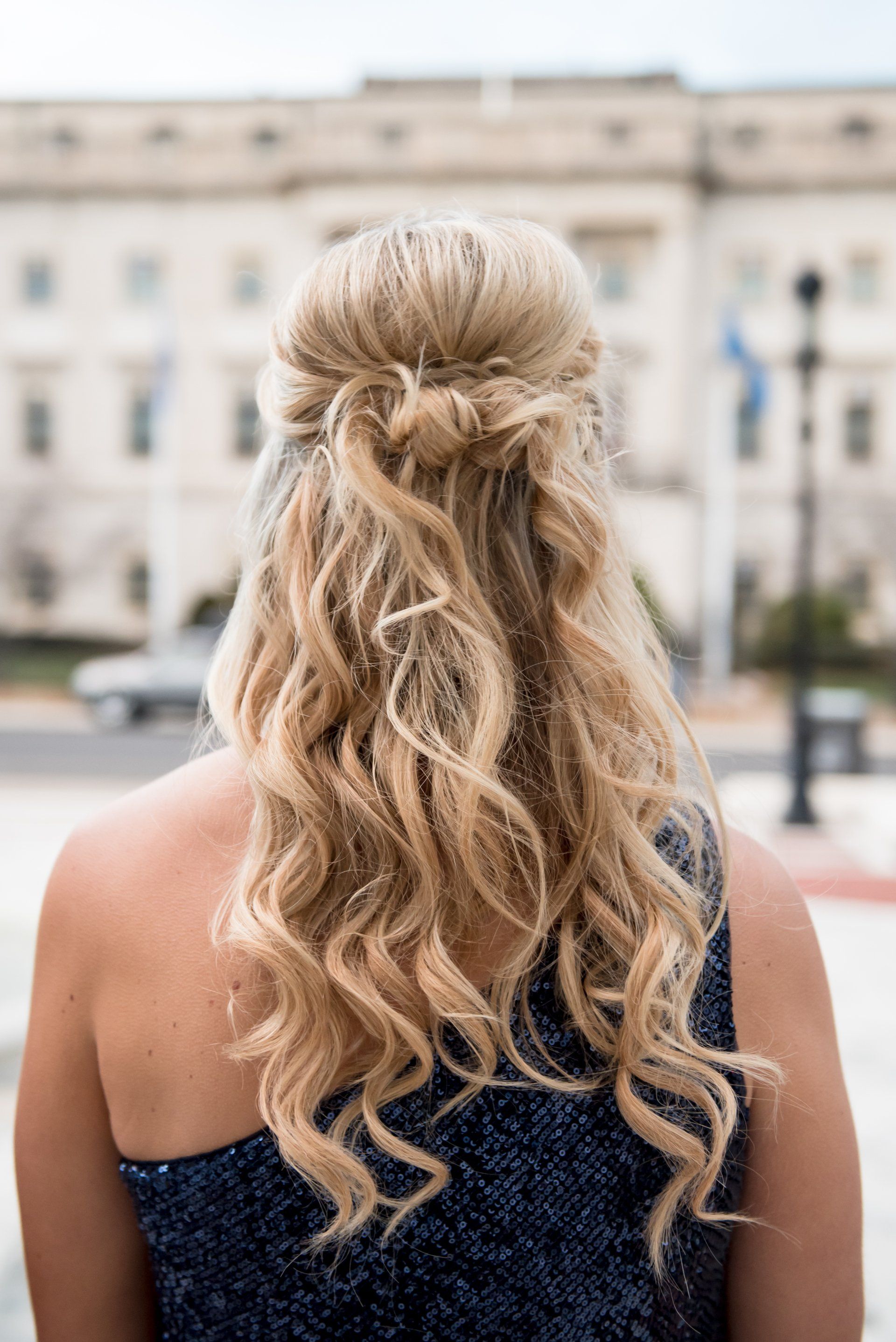 Back view of a woman with wavy blonde hair in an off-shoulder navy dress, standing outdoors near a building