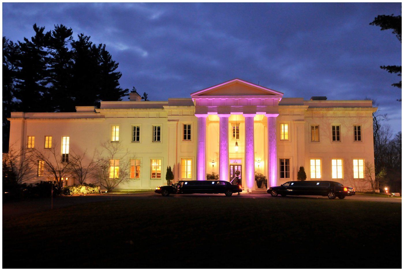 Illuminated White House at dusk with pink-lit columns and cars parked in front
