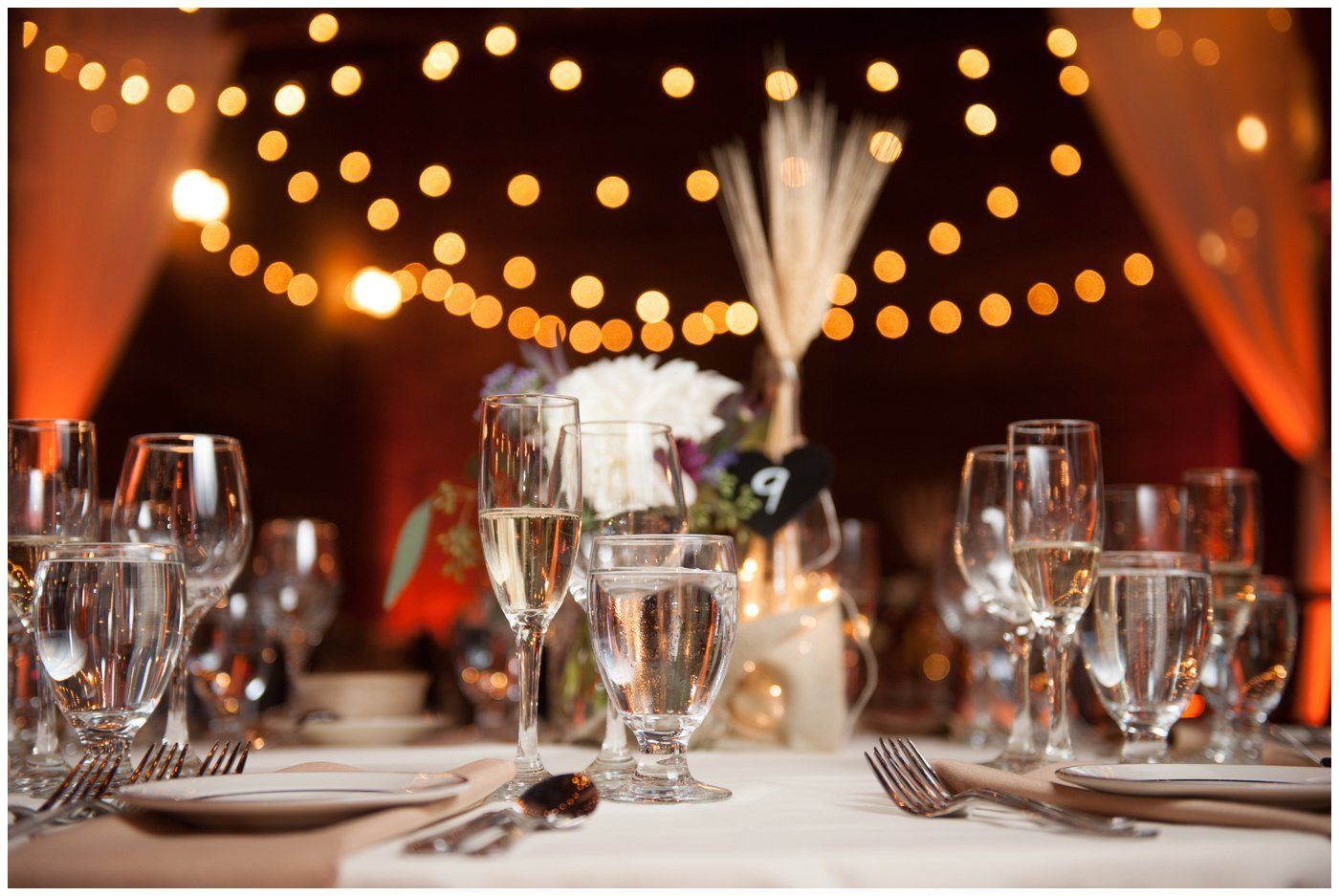 Elegant dinner table with wine glasses and silverware under warm string lights, softly blurred centerpiece behind.