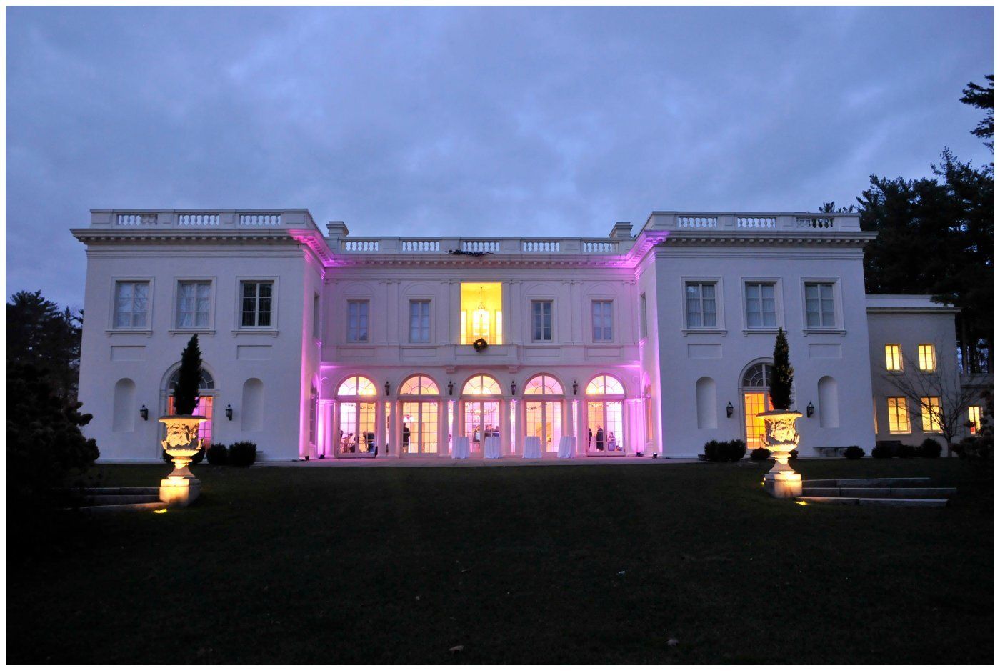 Grand white mansion lit with pink and yellow lights at dusk, viewed from a dark lawn