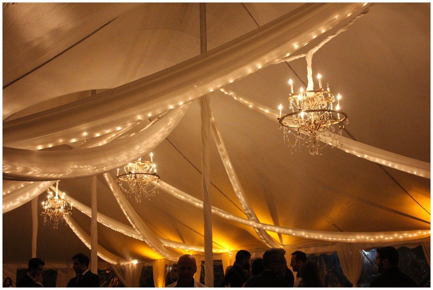 Elegant tented event with draped fabric, warm string lights, chandeliers, and a silhouetted crowd below