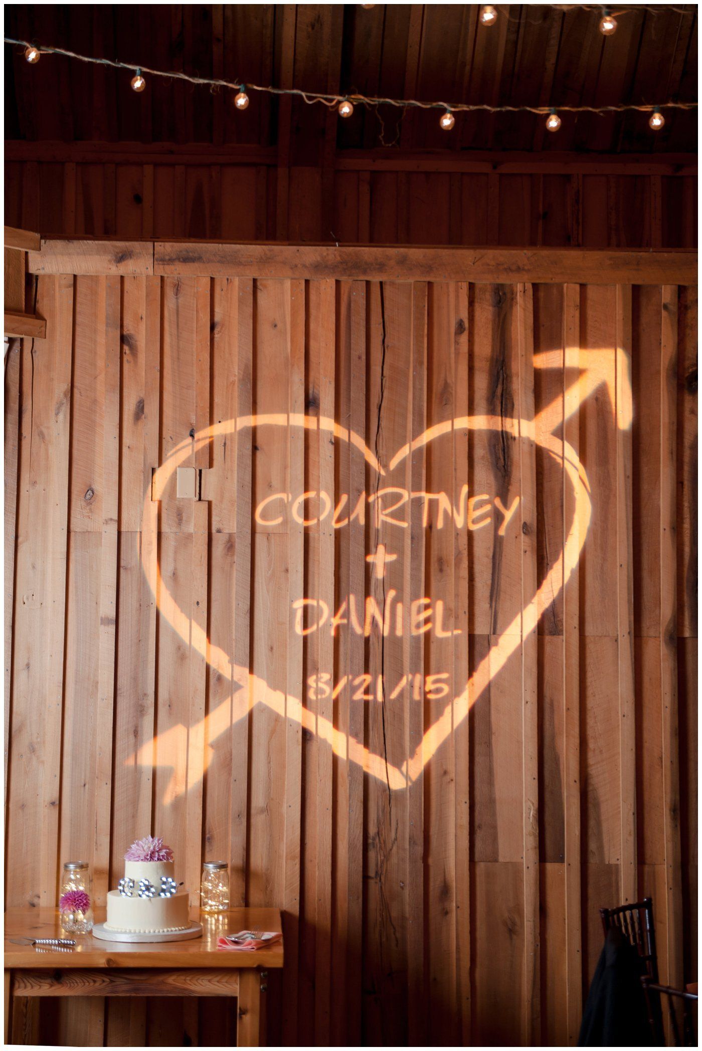 Neon heart and arrow sign on a wooden wall reads “Courtney + Daniel 8/2/15” under string lights.