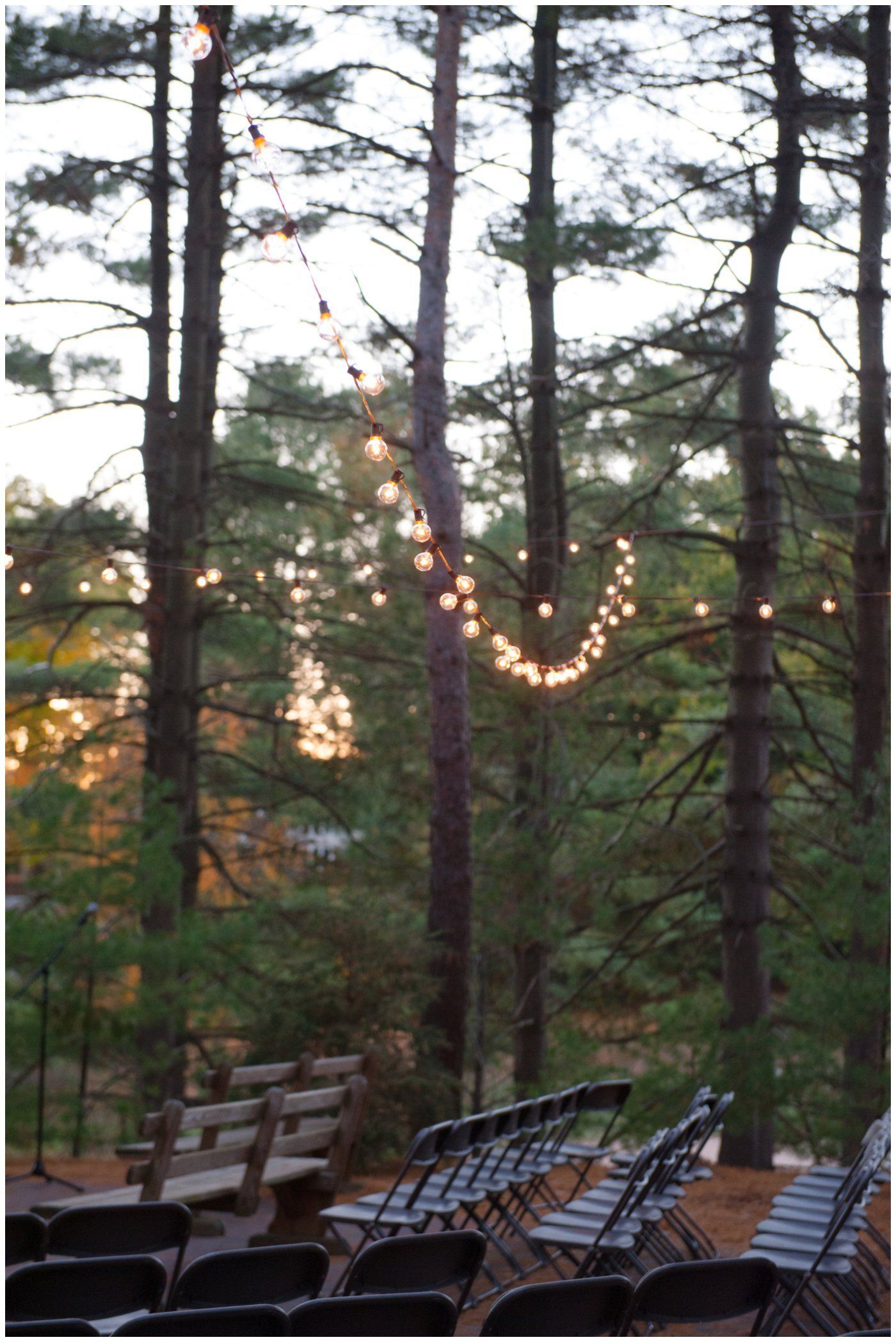 String lights hang between tall trees over empty chairs on a wooded patio at dusk.