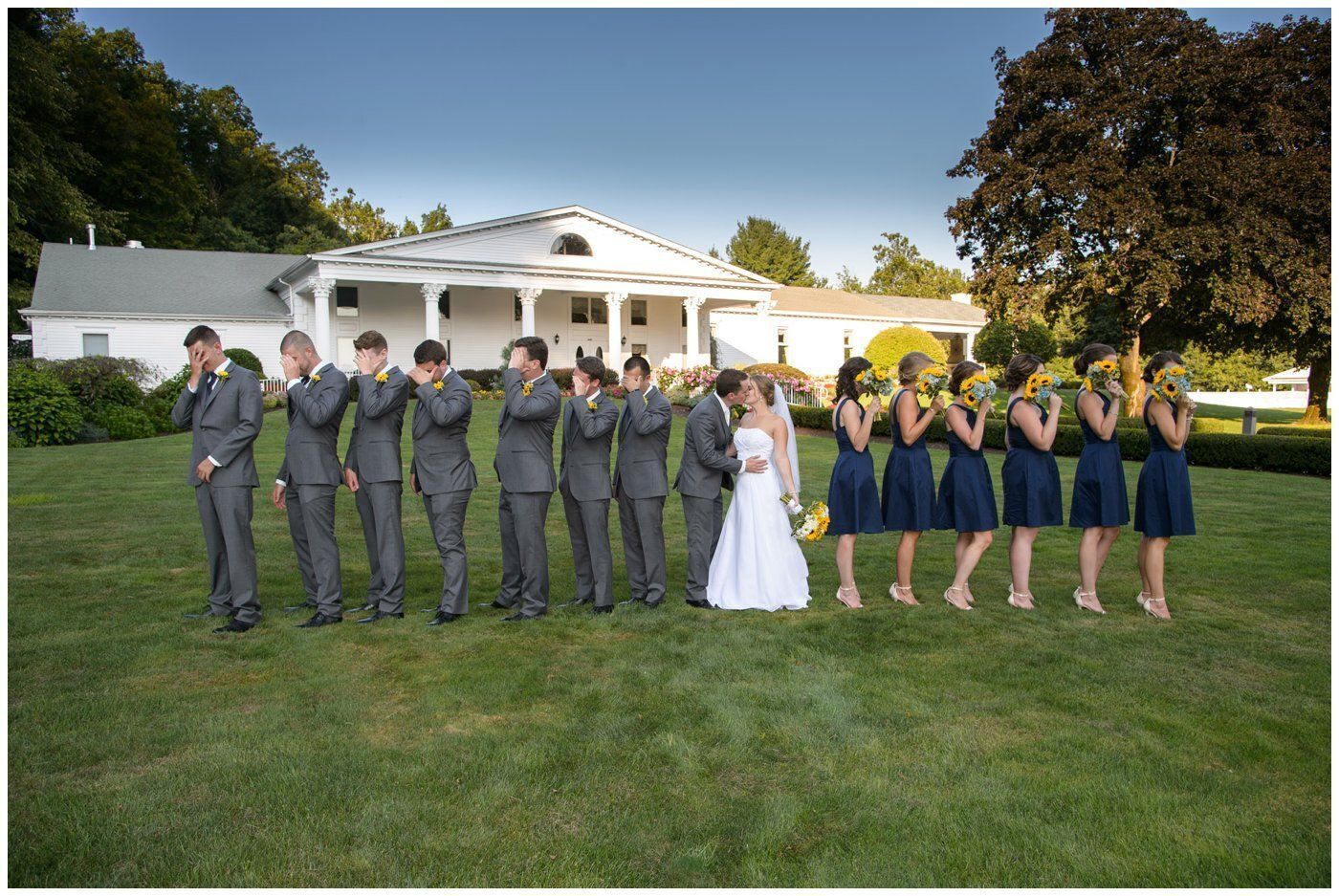 Wedding party poses on a lawn in front of a white mansion, with the bride and groom at center.