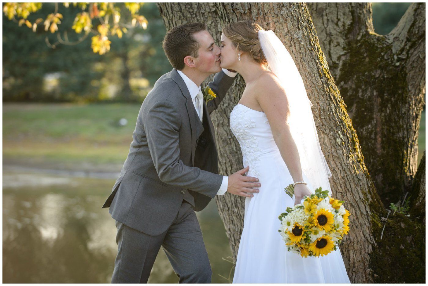 Bride and groom kissing beside a tree, with the bride holding a yellow bouquet outdoors.