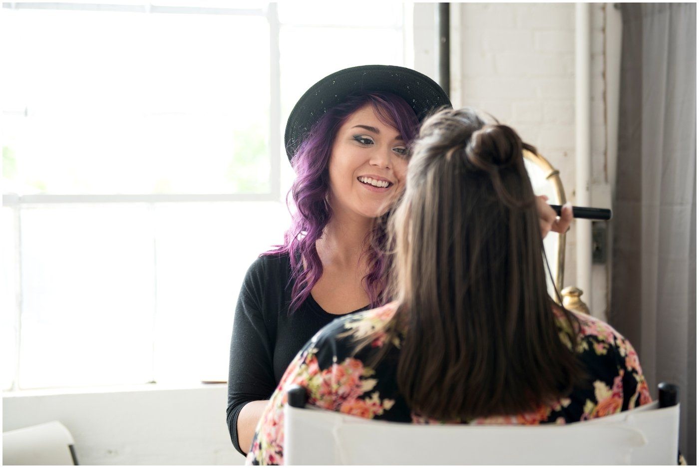 Two women talking in a bright room, one smiling in a black hat with purple hair