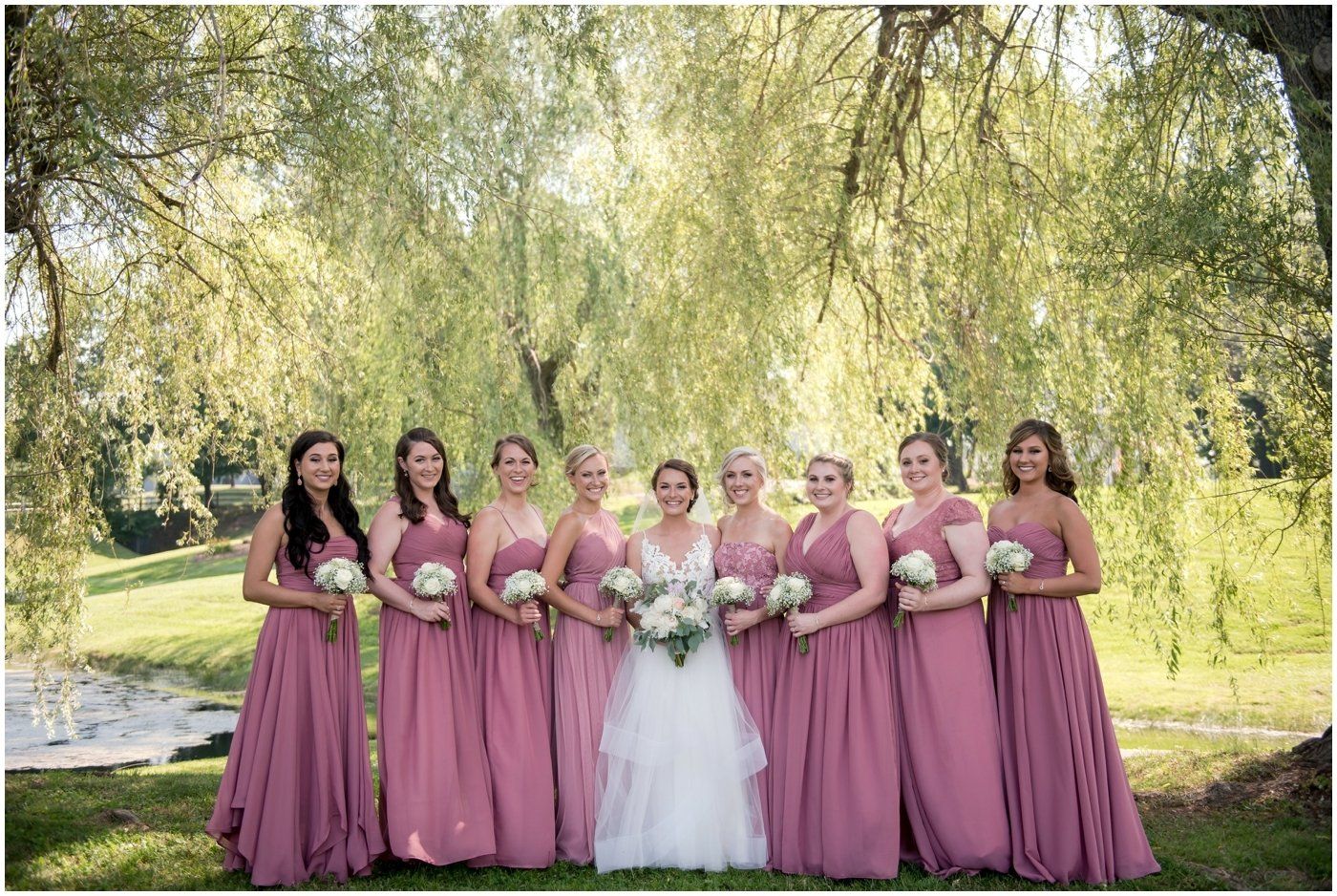 Bride with seven bridesmaids in mauve dresses holding bouquets outdoors under willow trees