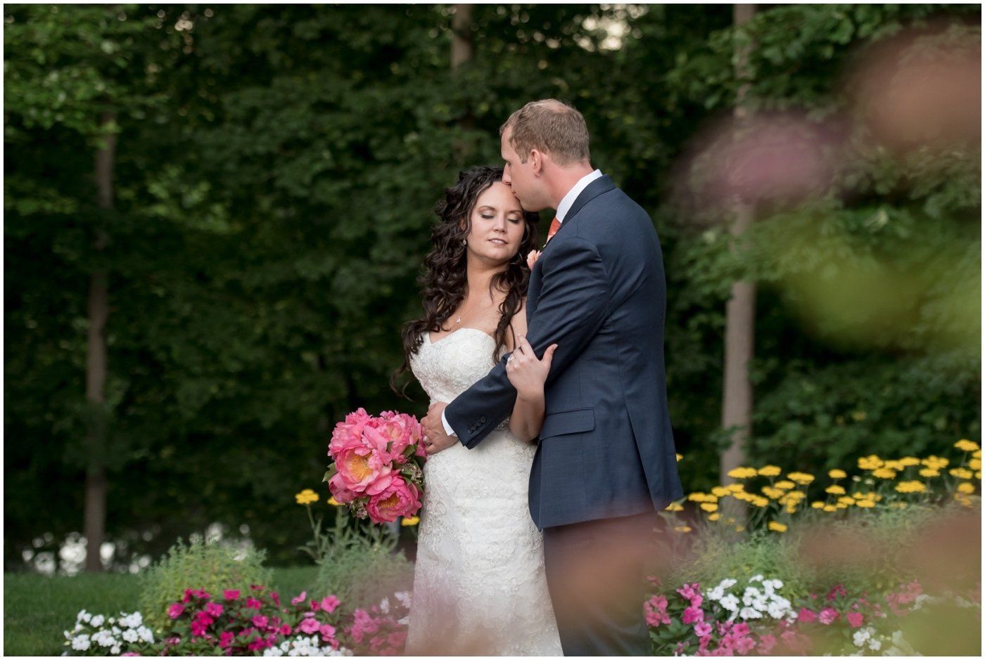 Bride and groom embracing in a flower garden, with pink bouquet and colorful blooms in the foreground