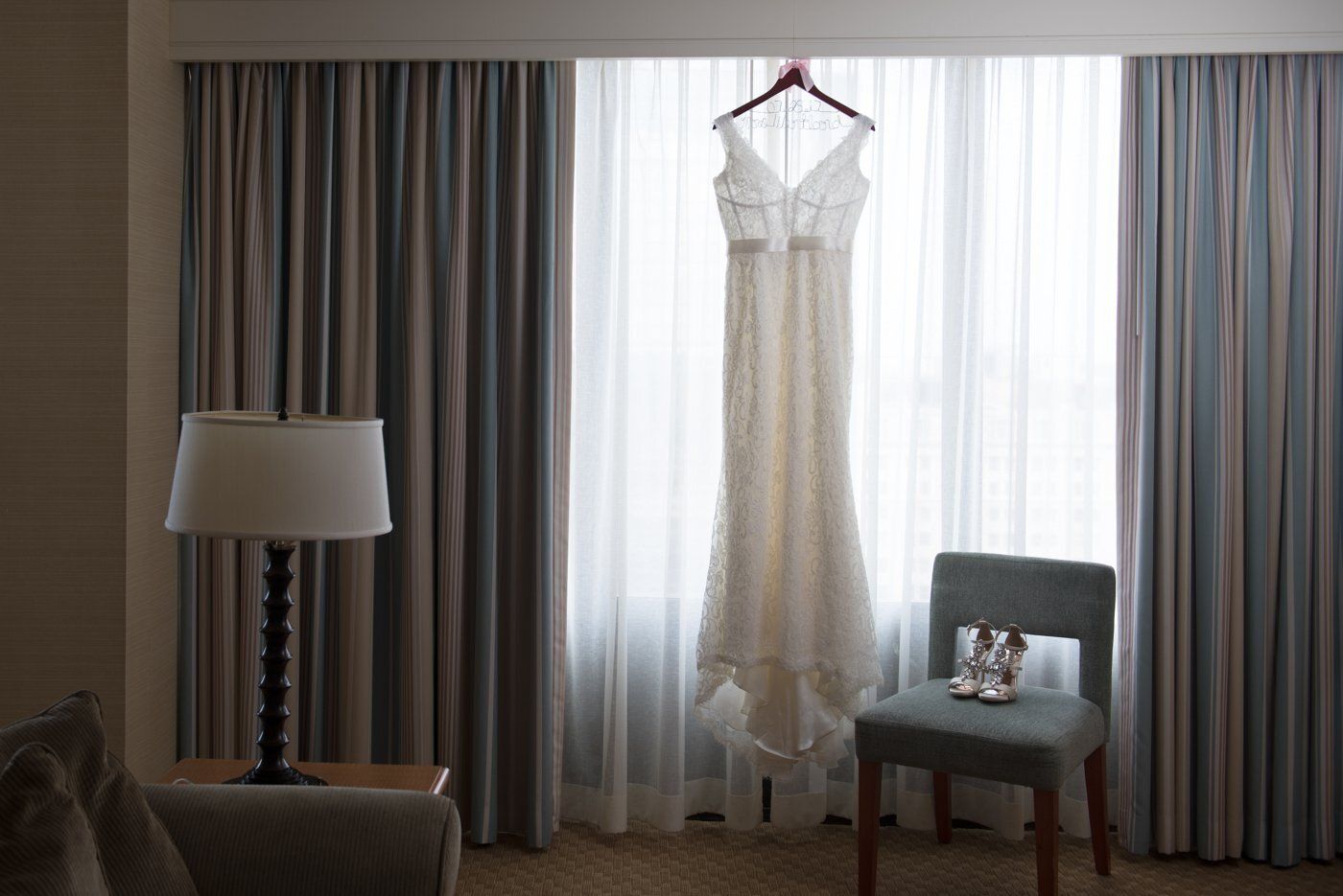 White lace dress hanging in a sunlit room beside curtains, lamp, and chair.