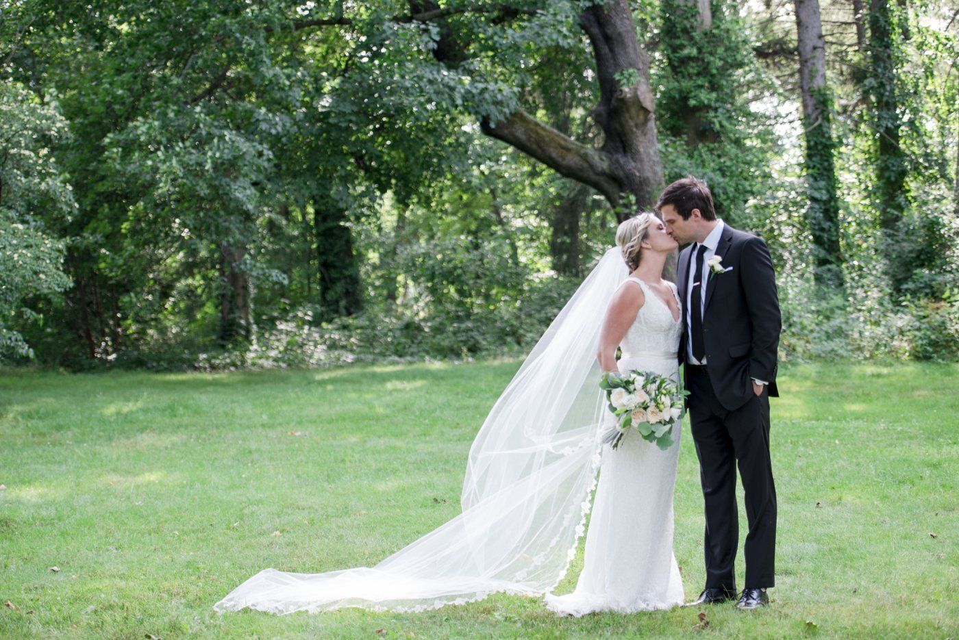 Bride and groom kissing in a green park, bride in white gown with long veil holding bouquet