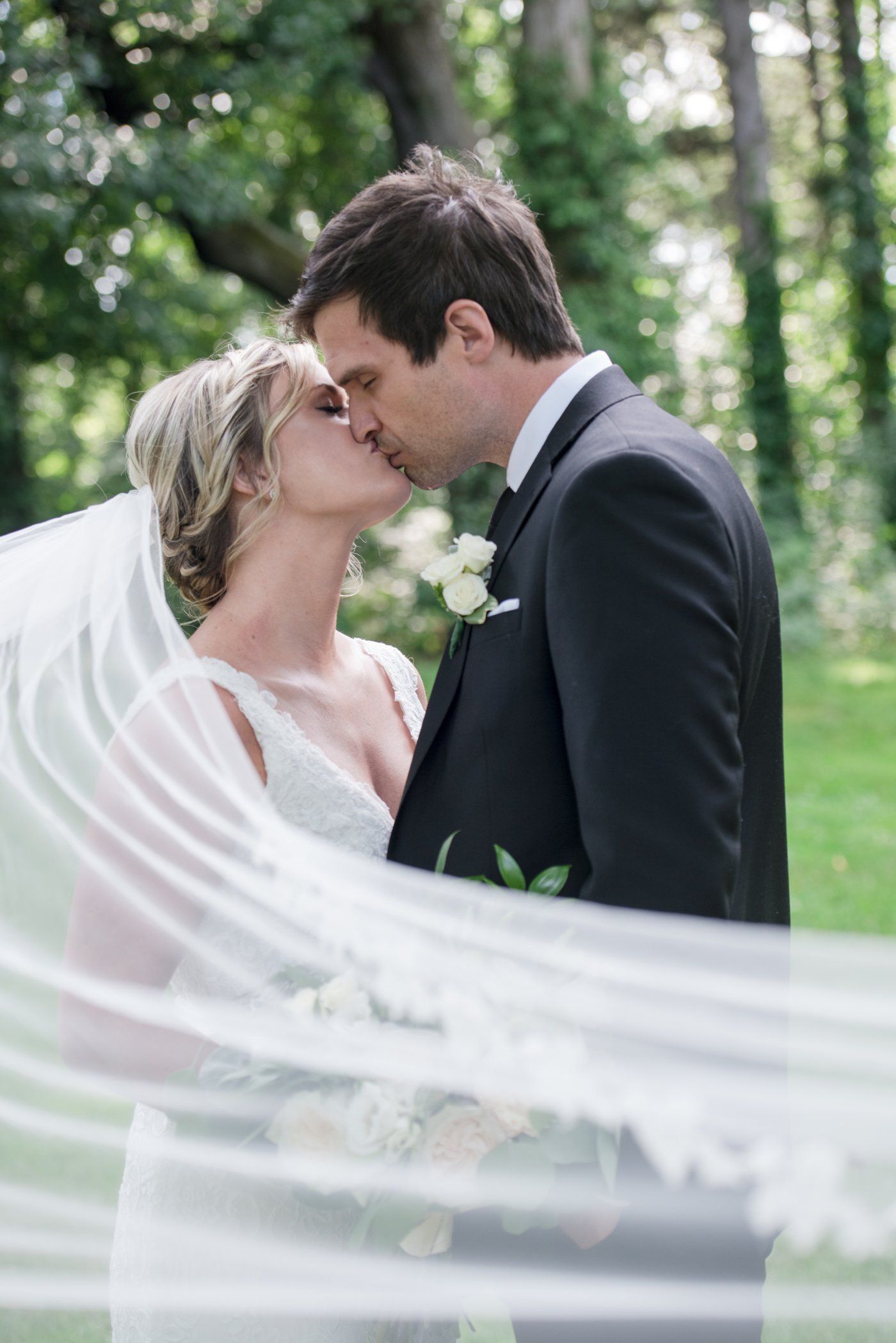 Bride and groom kiss in a wooded outdoor wedding scene, veil flowing in the foreground.