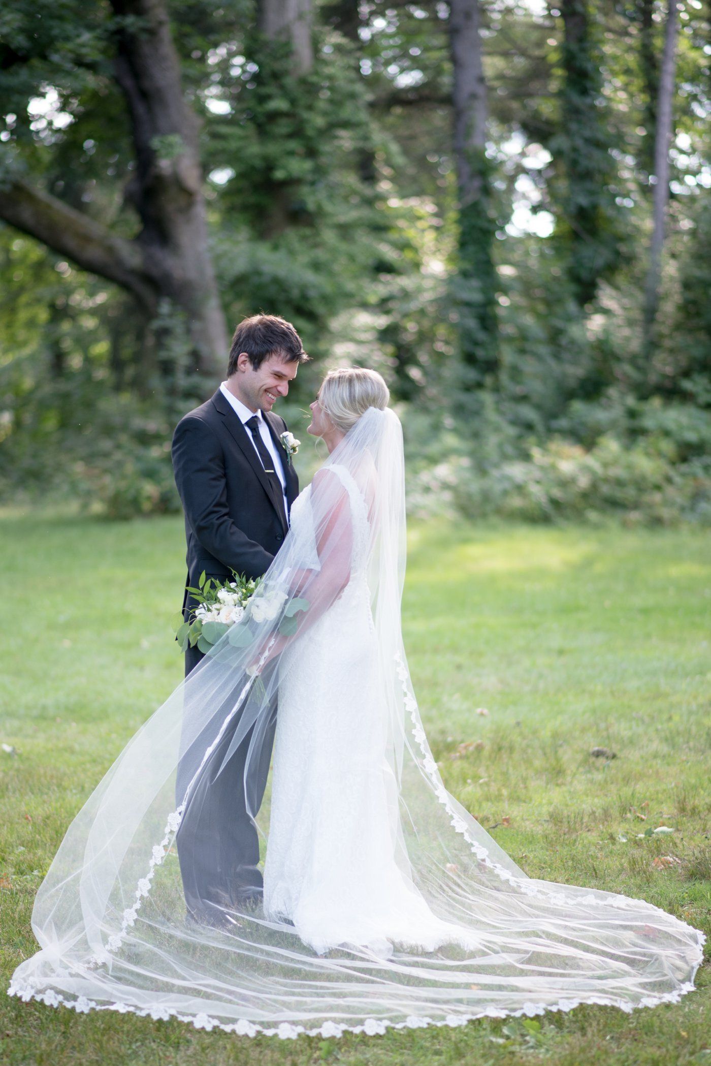 Bride and groom standing in a sunlit garden, sharing an intimate moment with a long veil spread on the grass