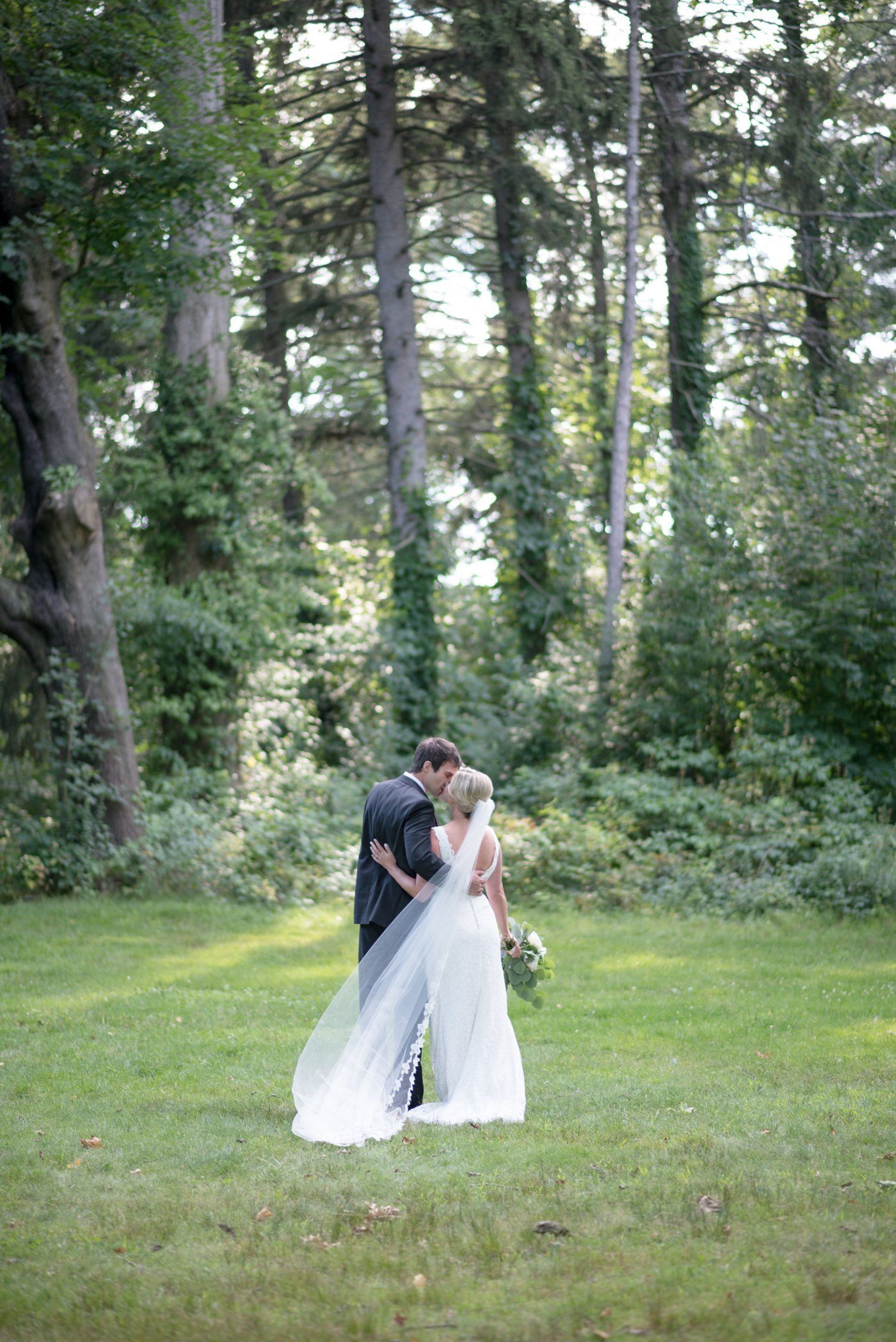 Bride and groom embracing in a green forest clearing, with the bride in a white gown and veil.