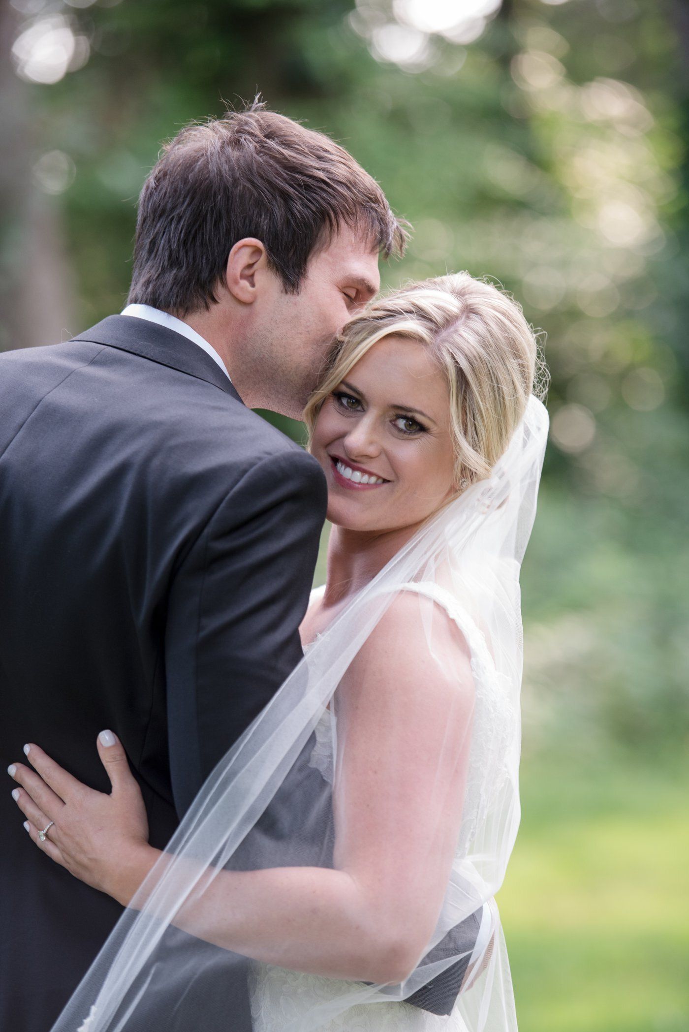 Bride smiling over groom’s shoulder outdoors, wearing a veil and white dress