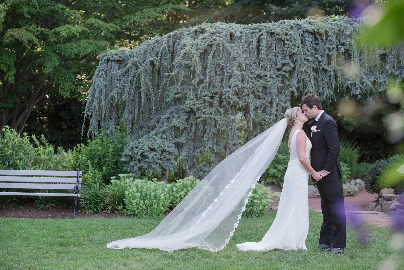 Bride and groom kissing in a garden, with the bride’s veil flowing across the grass