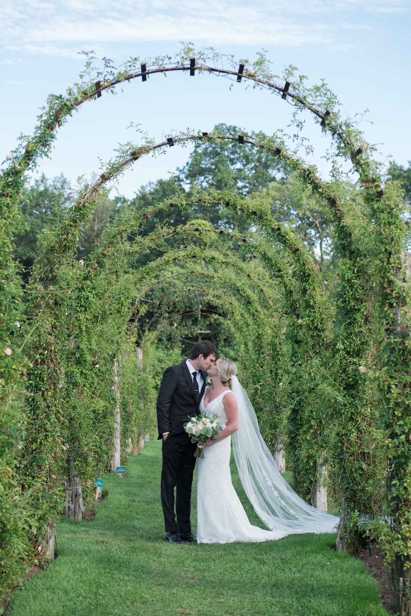 Bride and groom kissing beneath a green garden arch tunnel outdoors