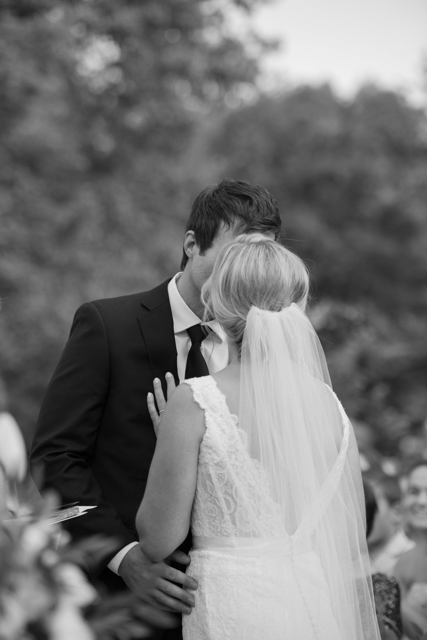 Wedding couple embracing outdoors, with the bride in a veil and groom in a dark suit.