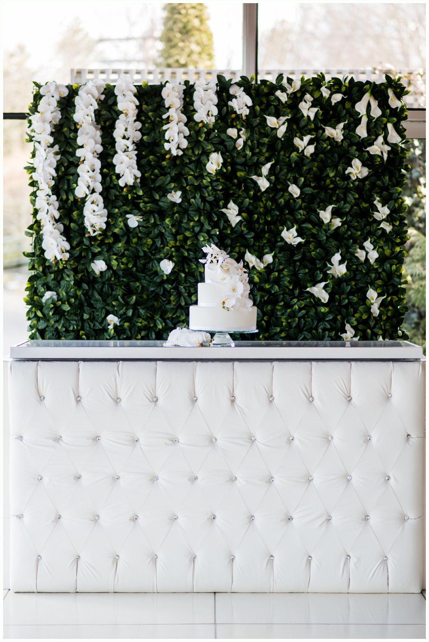 White tufted display table with a green floral wall and hanging white flowers behind it