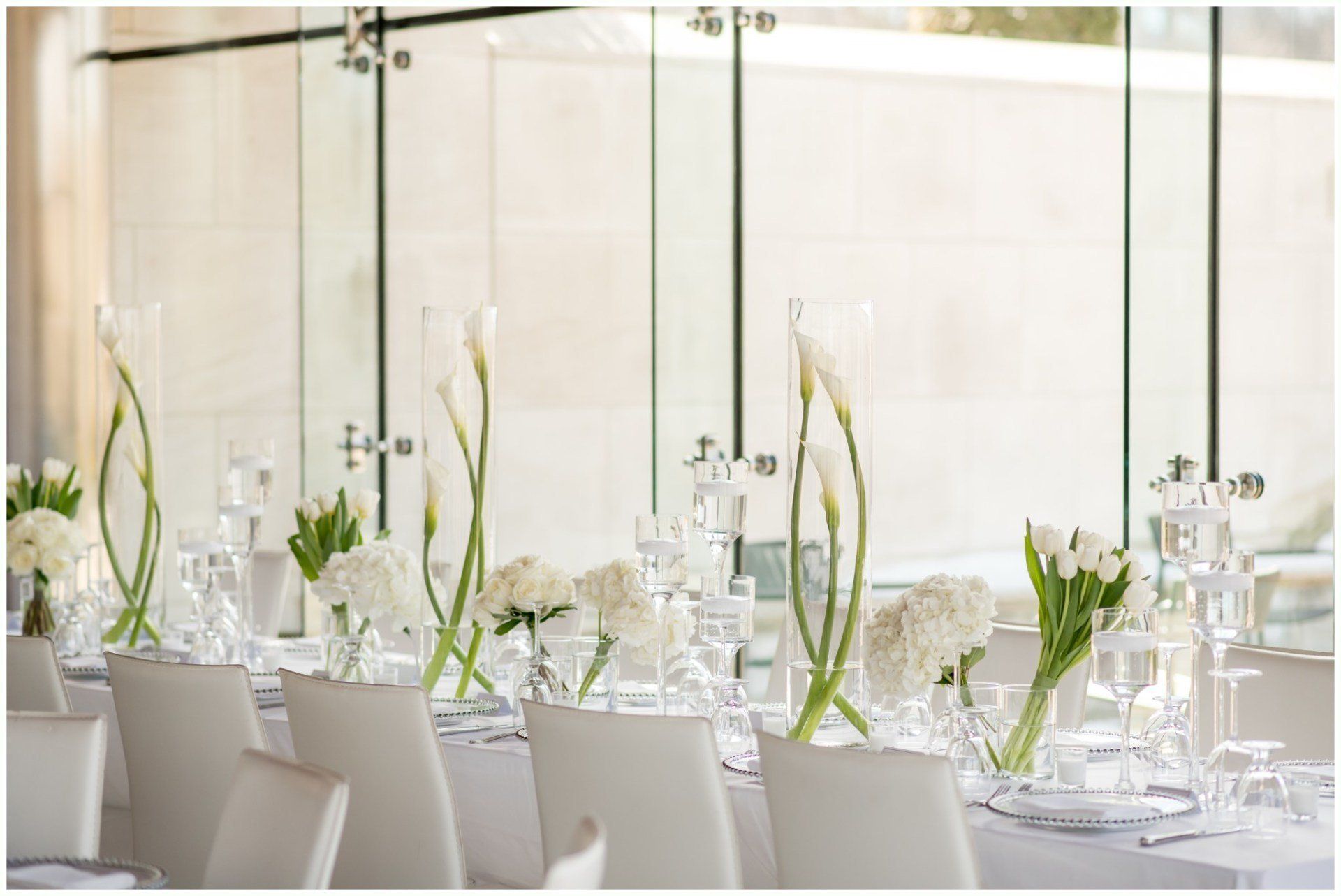 Elegant white dining table set with glassware and tall green floral centerpieces in a bright room