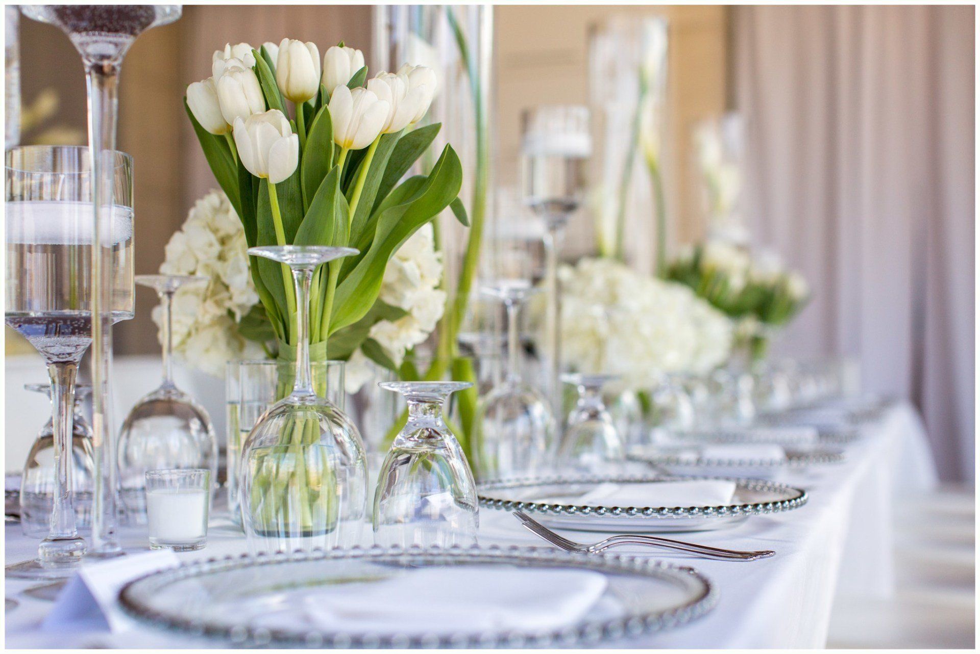 Elegant table setting with white tulips, glassware, and silver chargers on a white-draped banquet table