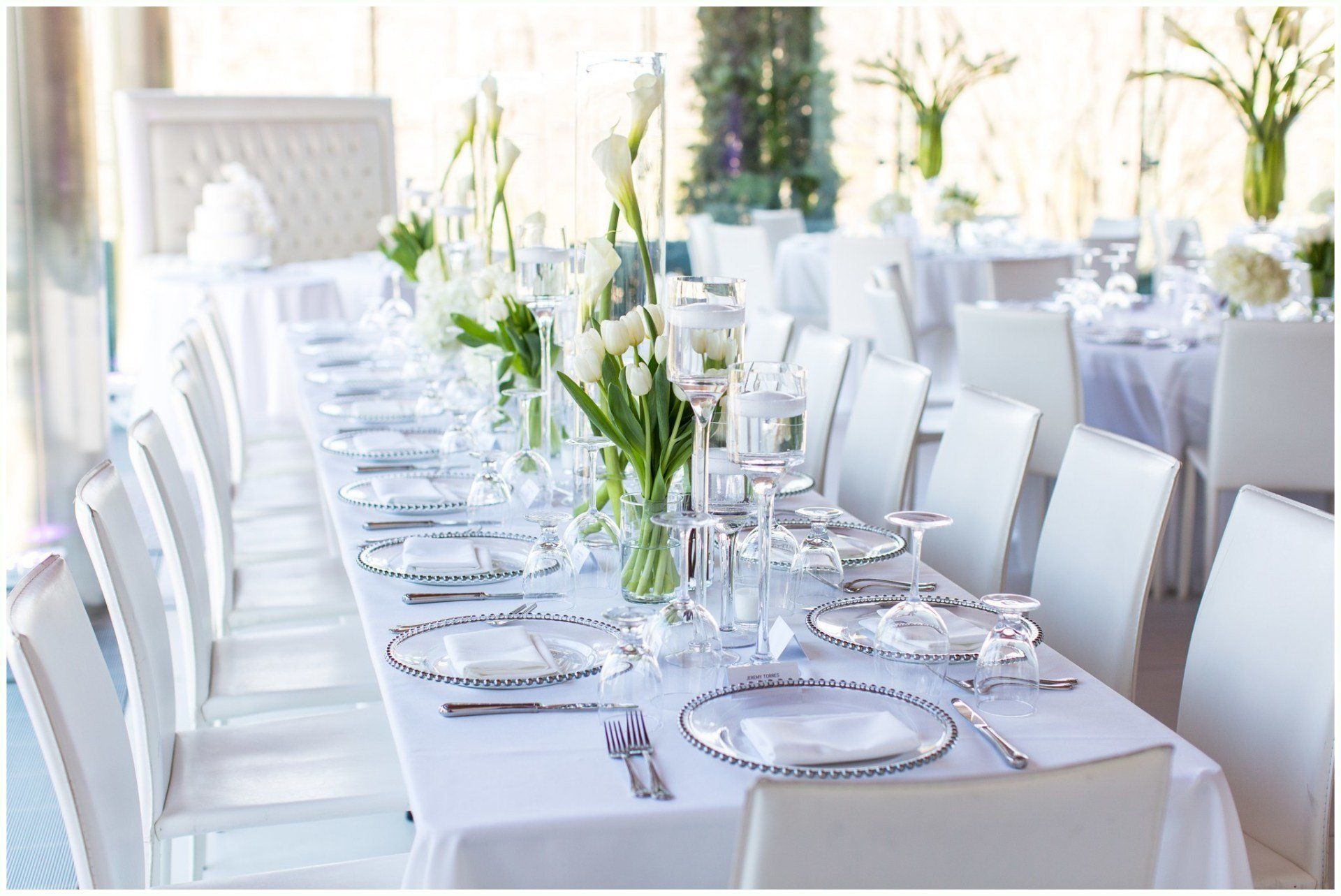 White-clothed banquet table set with plates, glasses, and white floral centerpieces in a bright event hall