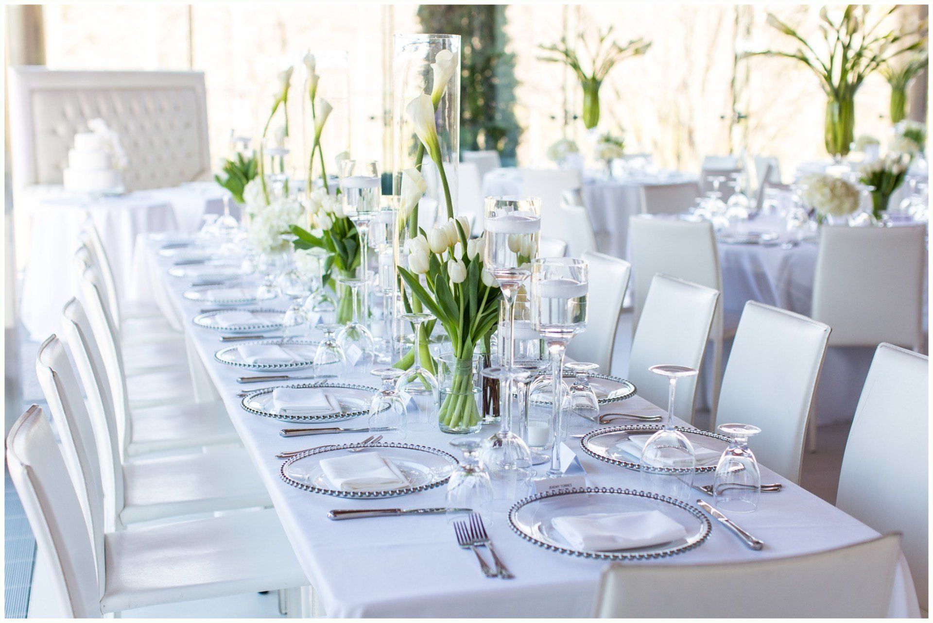 Elegant white banquet table set with glassware, plates, and floral centerpieces in a bright room