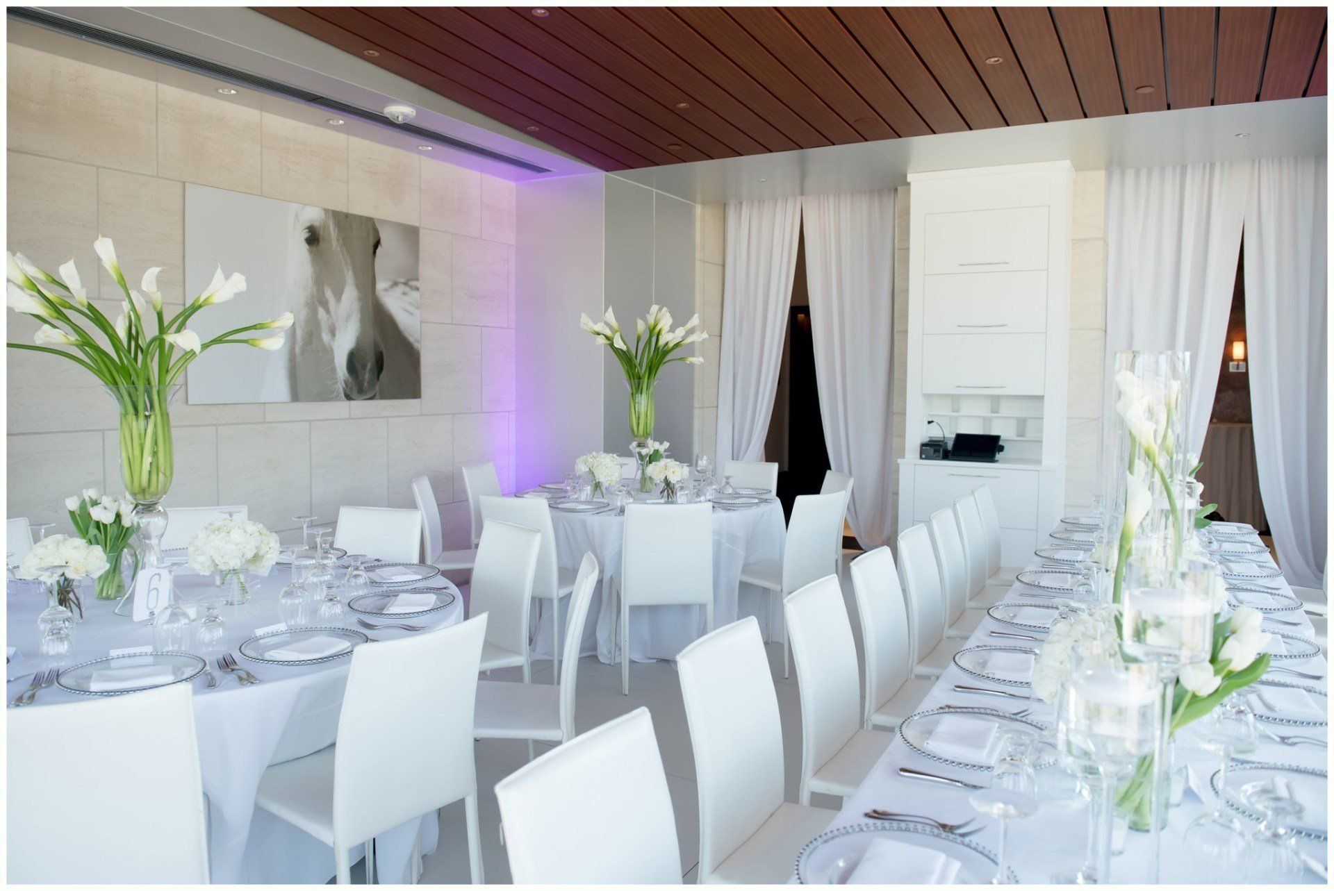 Elegant banquet hall with white-dressed tables, white chairs, floral centerpieces, and soft purple lighting