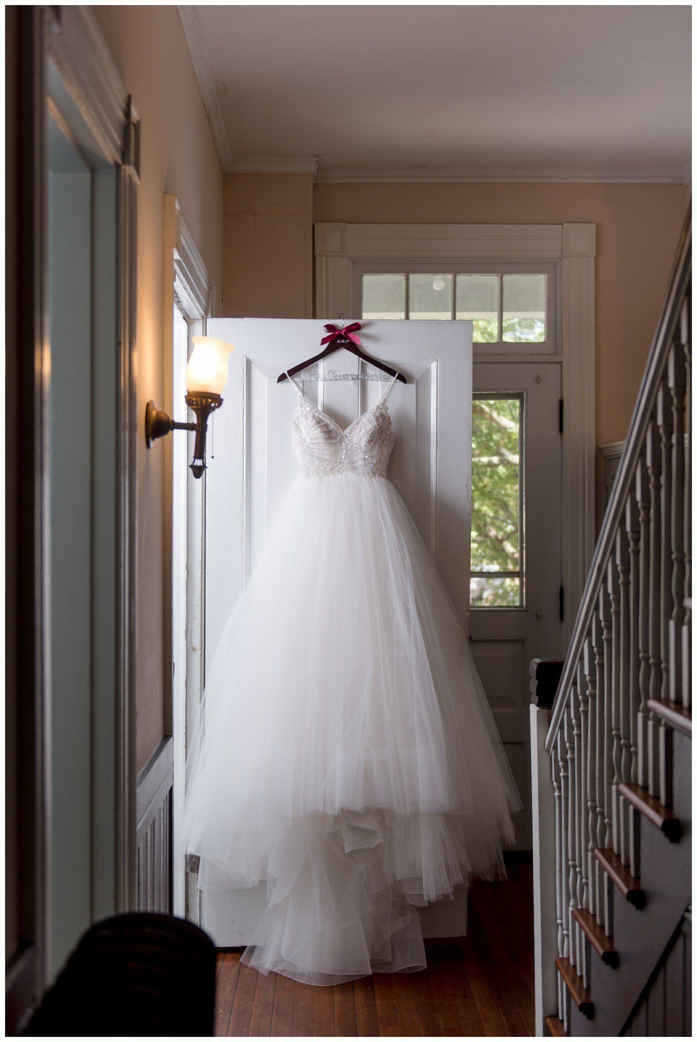 Wedding dress hanging in a hallway by a front door, with a dark red sash on the hanger