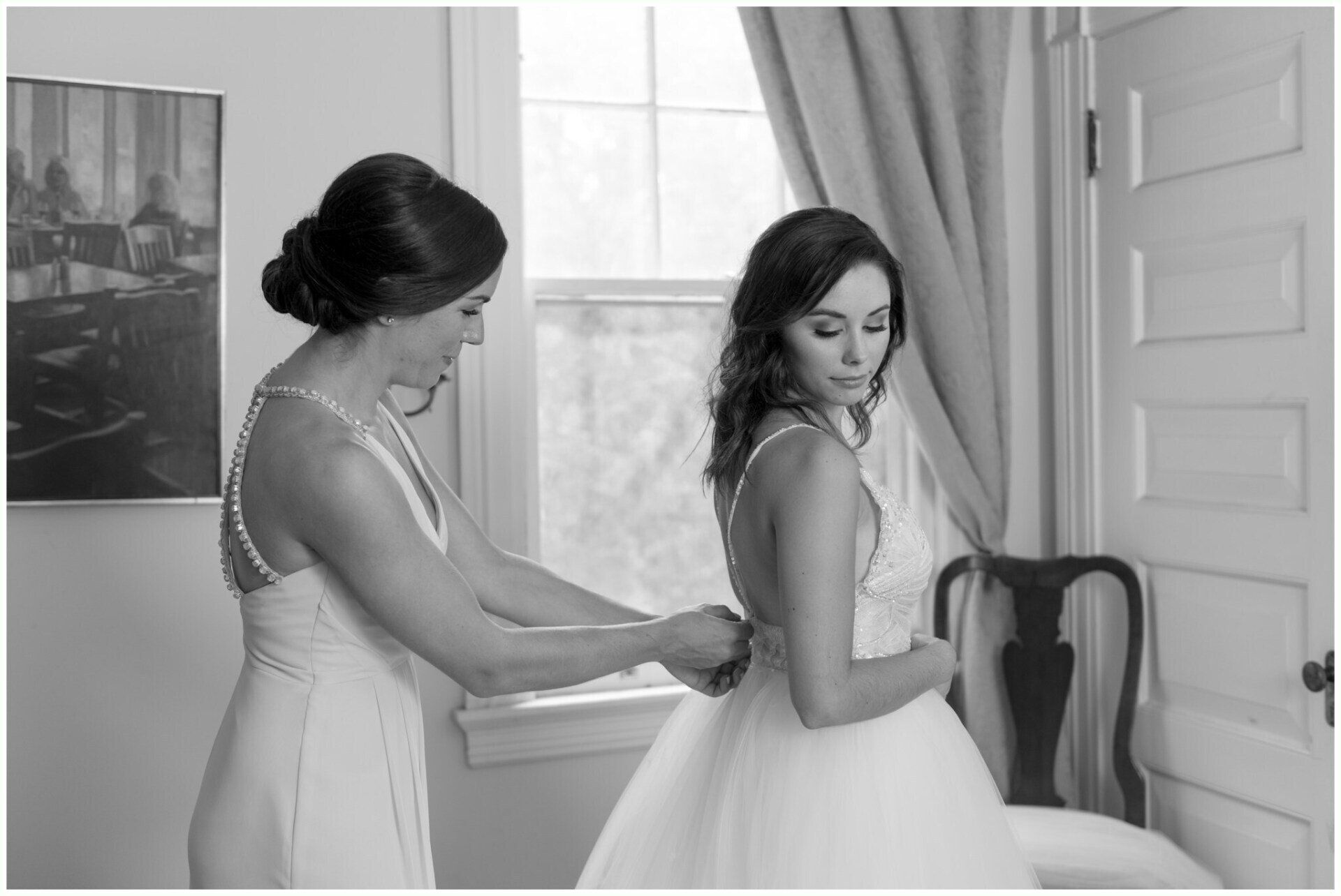 Two women in white dresses adjusting a gown in a bright room near a window