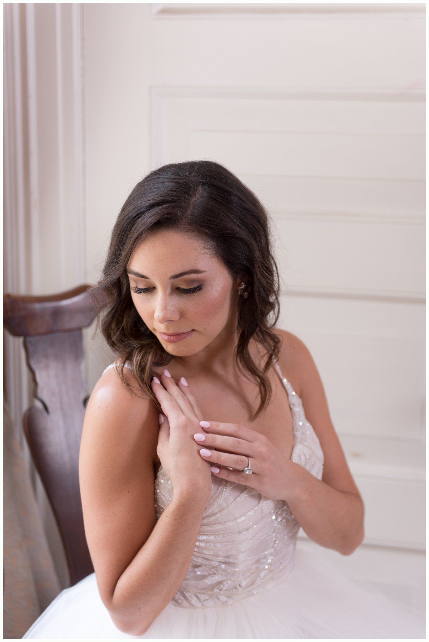 Bride in a white lace dress looking down by a window, hands crossed over her chest